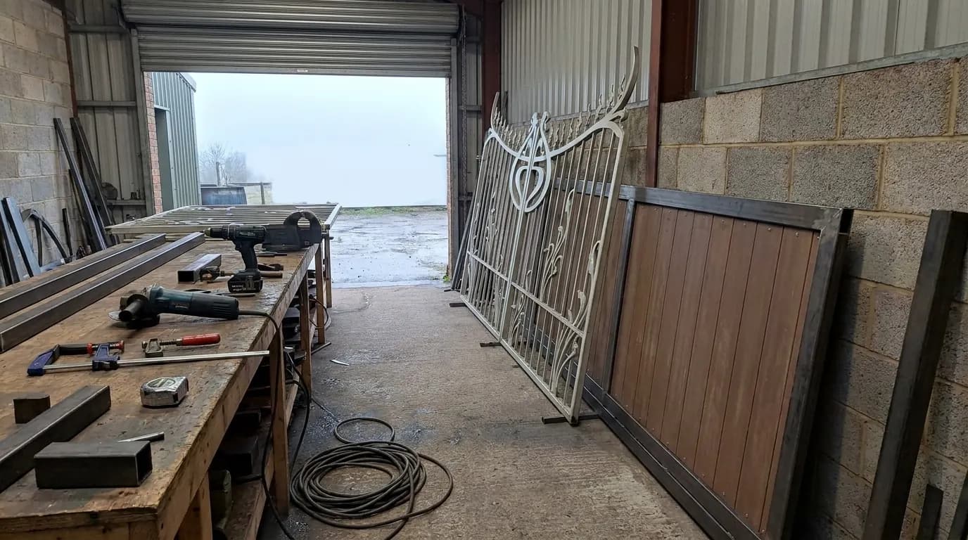 Steel gate sections leaning against a wall in a small workshop yard with hand tools on a bench
