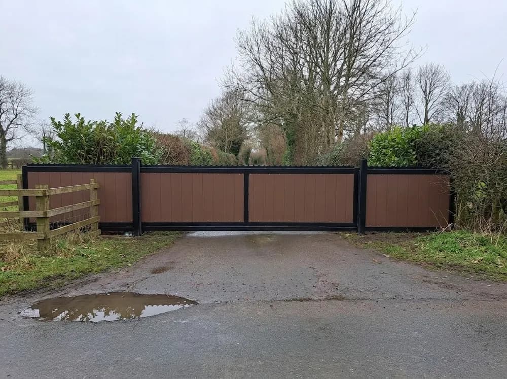 A wide brown composite board gate with a black steel frame closed across a rural lane entrance