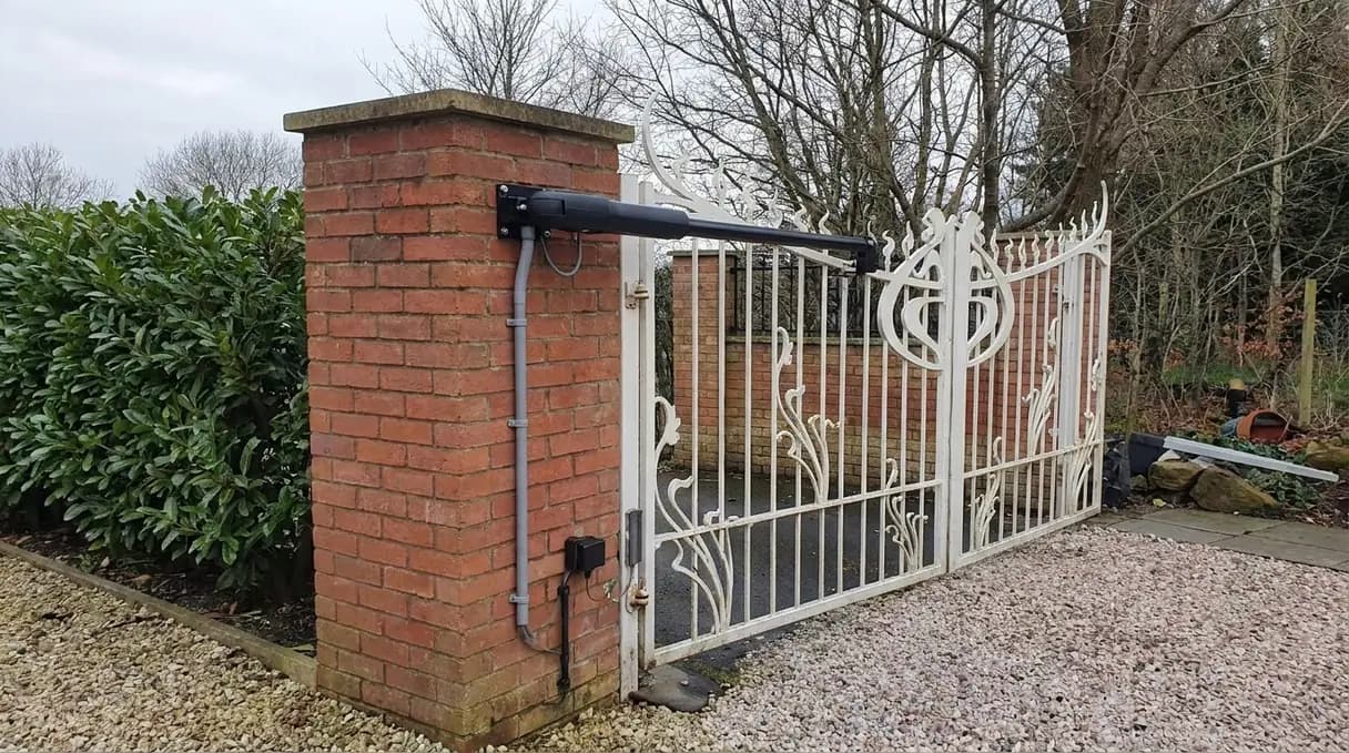 An electric motor arm fitted to a white painted iron driveway gate at a red brick pillar, with cabling clipped neatly in conduit