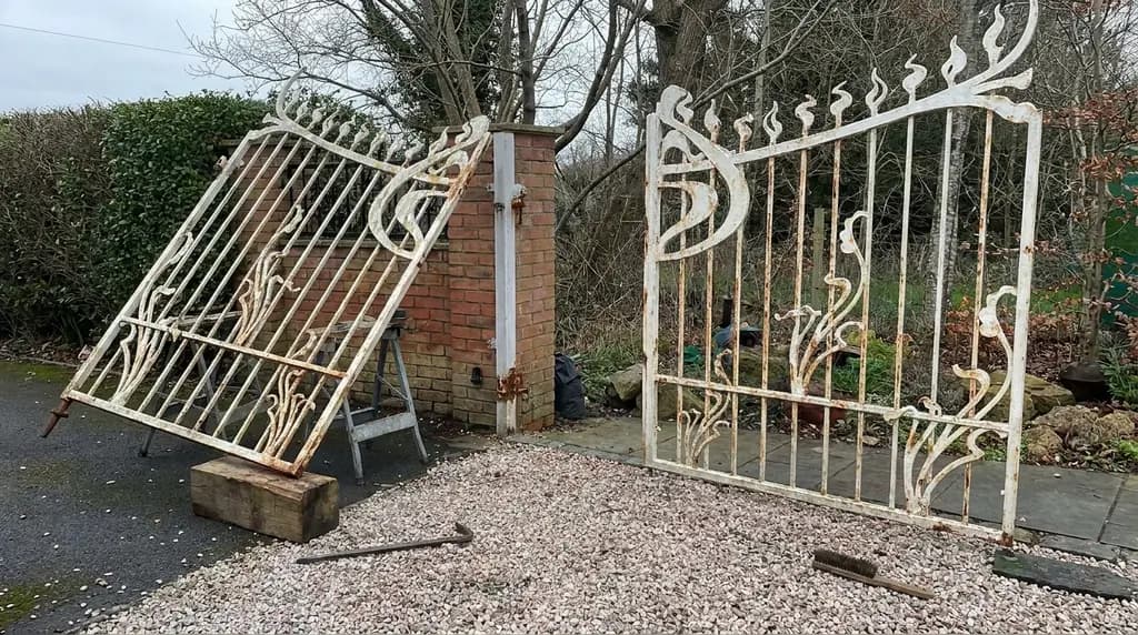 An older wrought iron gate with flaking paint and a sagging leaf lifted off its hinge and propped during repair